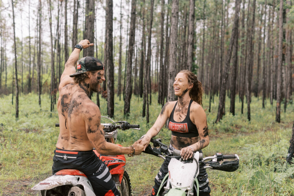 couple laugh at each other sitting on dirt bikes in the woods during their engagement photo shoot