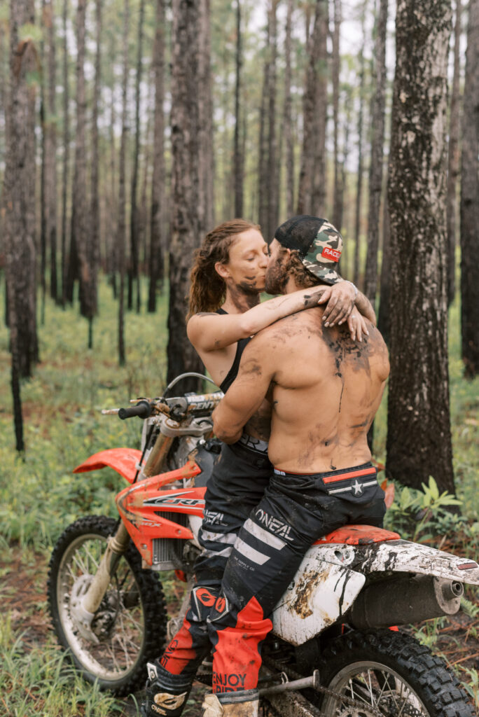 girlfriend sits on her boyfriends dirt bike and they kiss while covered in mud in the middle of the woods