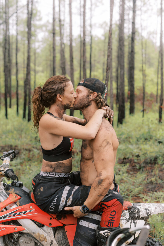 couple kissing while sitting on a red dirt bike in the middle of the woods covered in mud