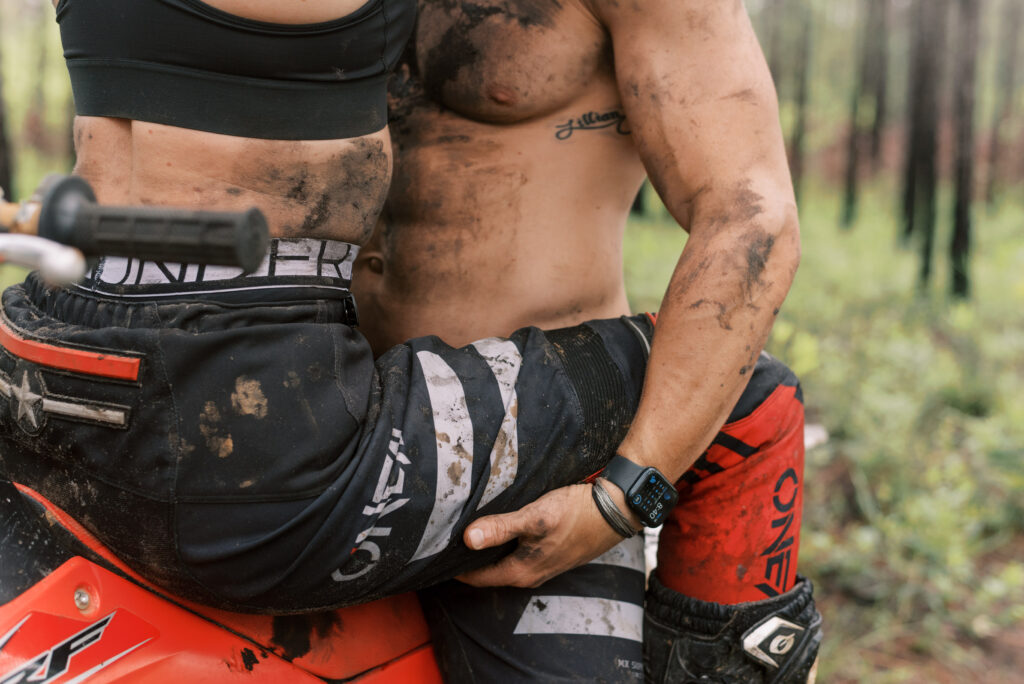 boyfriend holds his girlfriend on his lap sitting on his red dirt bike in the woods