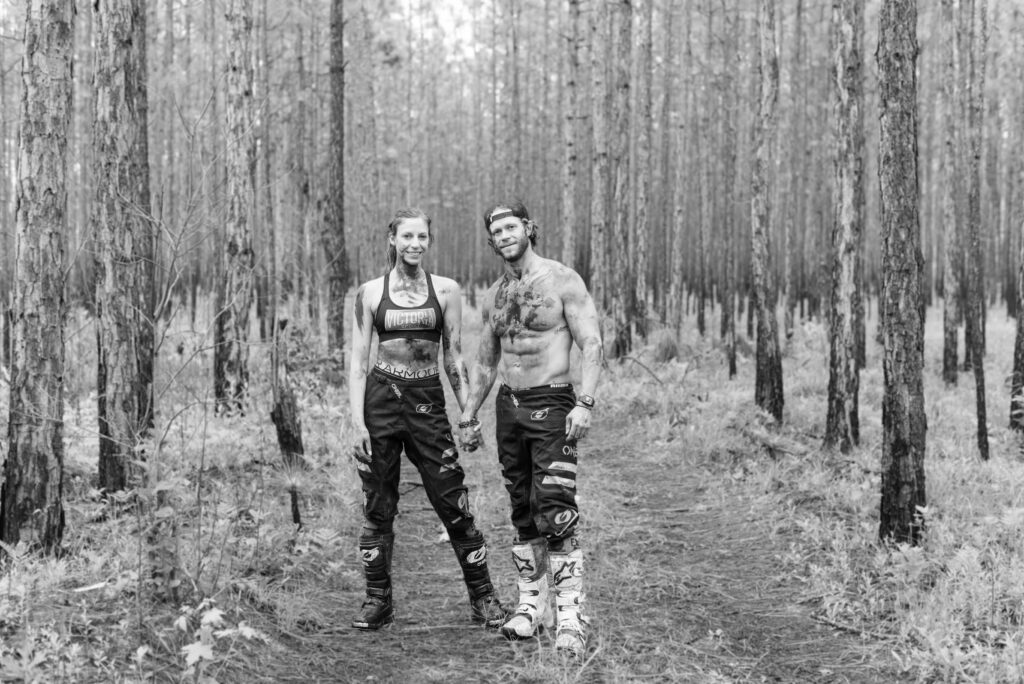 Black and white image of a couple holding hands in the woods smiling at the camera wearing only oneil dirt bike pants and covered in mud