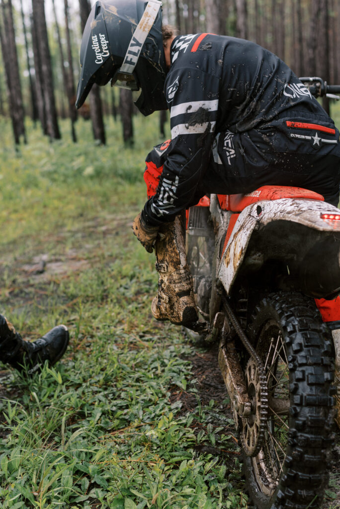 Man fixing his dirt bike boot while sitting on the bike wearing black and red Oneil bike clothes and a black helmet