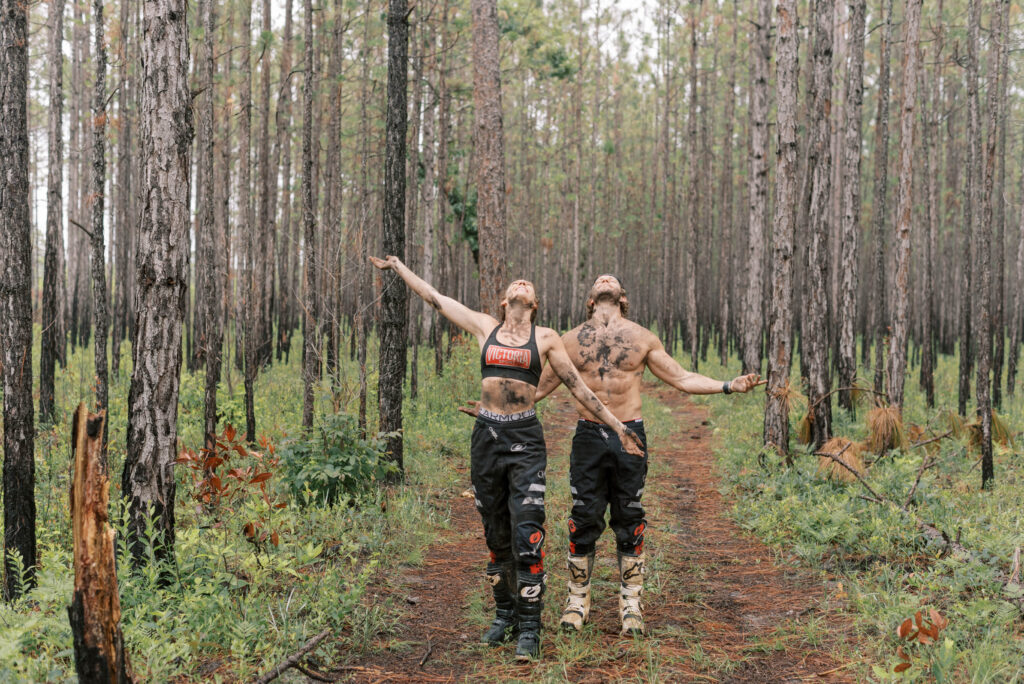 Couple stand with their faces upturned towards the rain and their arms out. They are covered in mud and standing in the middle of the woods