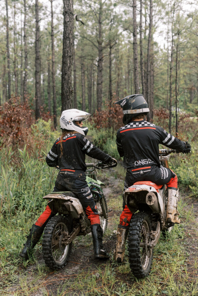 back view of two dirt bikers talking before riding off on a forest trail in the mud