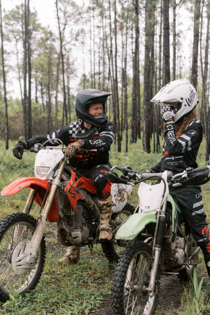 Two dirt bikers sit on their bikes beside each other, the man smiling at his partner as she fixes her goggles