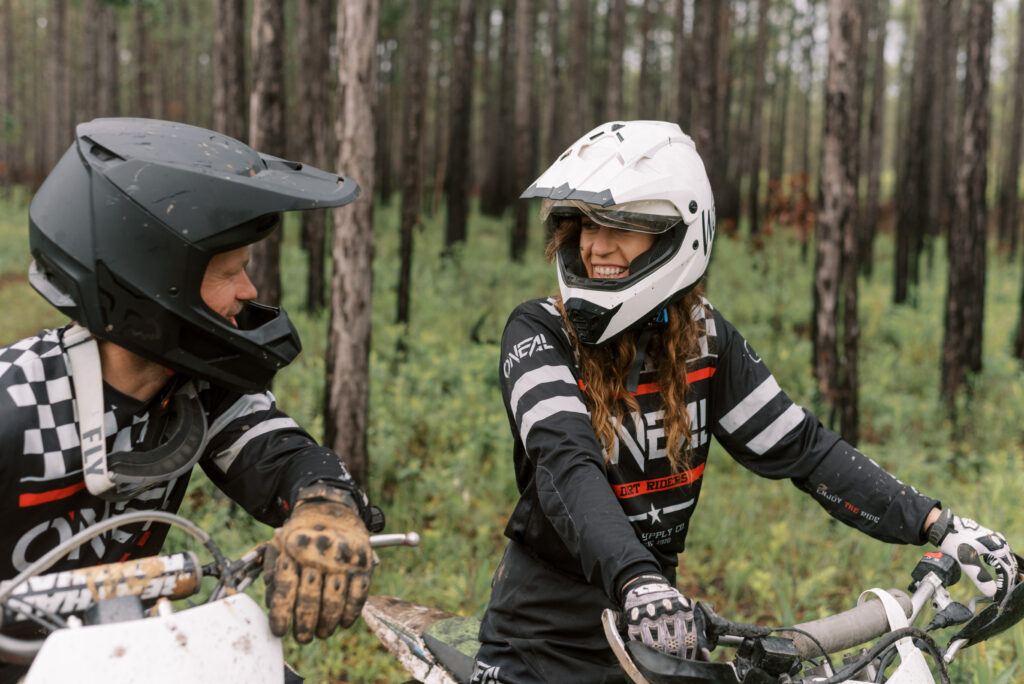 Woman smiles at her partner wearing a white helmet and sitting on her dirtbike