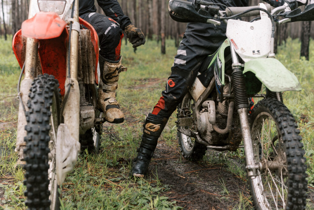 view of dirt bike riders legs and boots as they sit on their bikes and prepare to ride through the mud