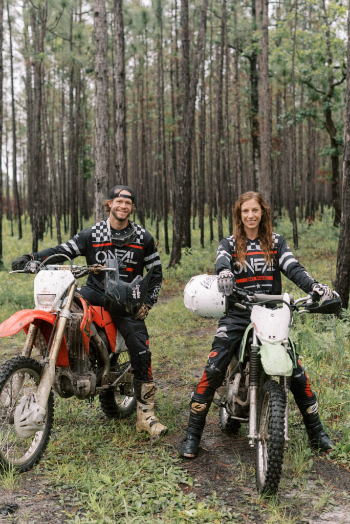 A couple smiles at the camera and sits on their dirt bikes in the woods