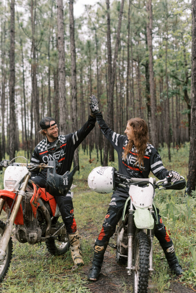 Couple high-fives while sitting on their dirt bikes in the woods. He has a black hat on and she has her hair down