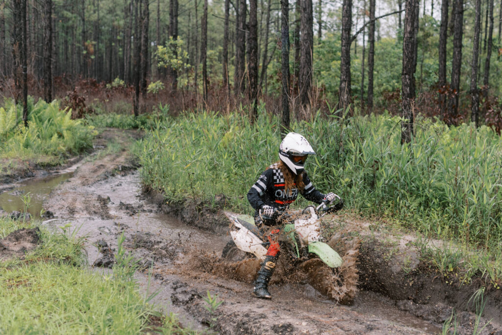 Woman in a white helmet and black oneil riding gear on a dirt bike riding through a splashing mud pit in the woods