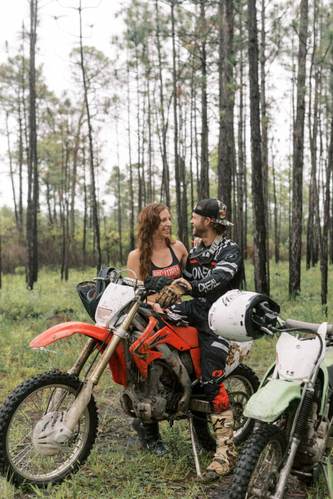 couple smile at each other and stand together while he sits on his dirt bike in the woods. 