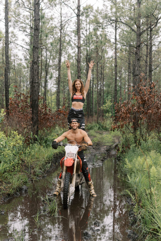 Man sits on his dirt bike and smiles at the camera while his girlfriend stands on the back of the bike and throws her hands in the air