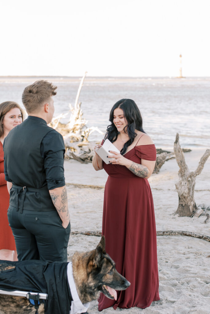 Bride in a red dress reads her vows on the beach with driftwood in the background